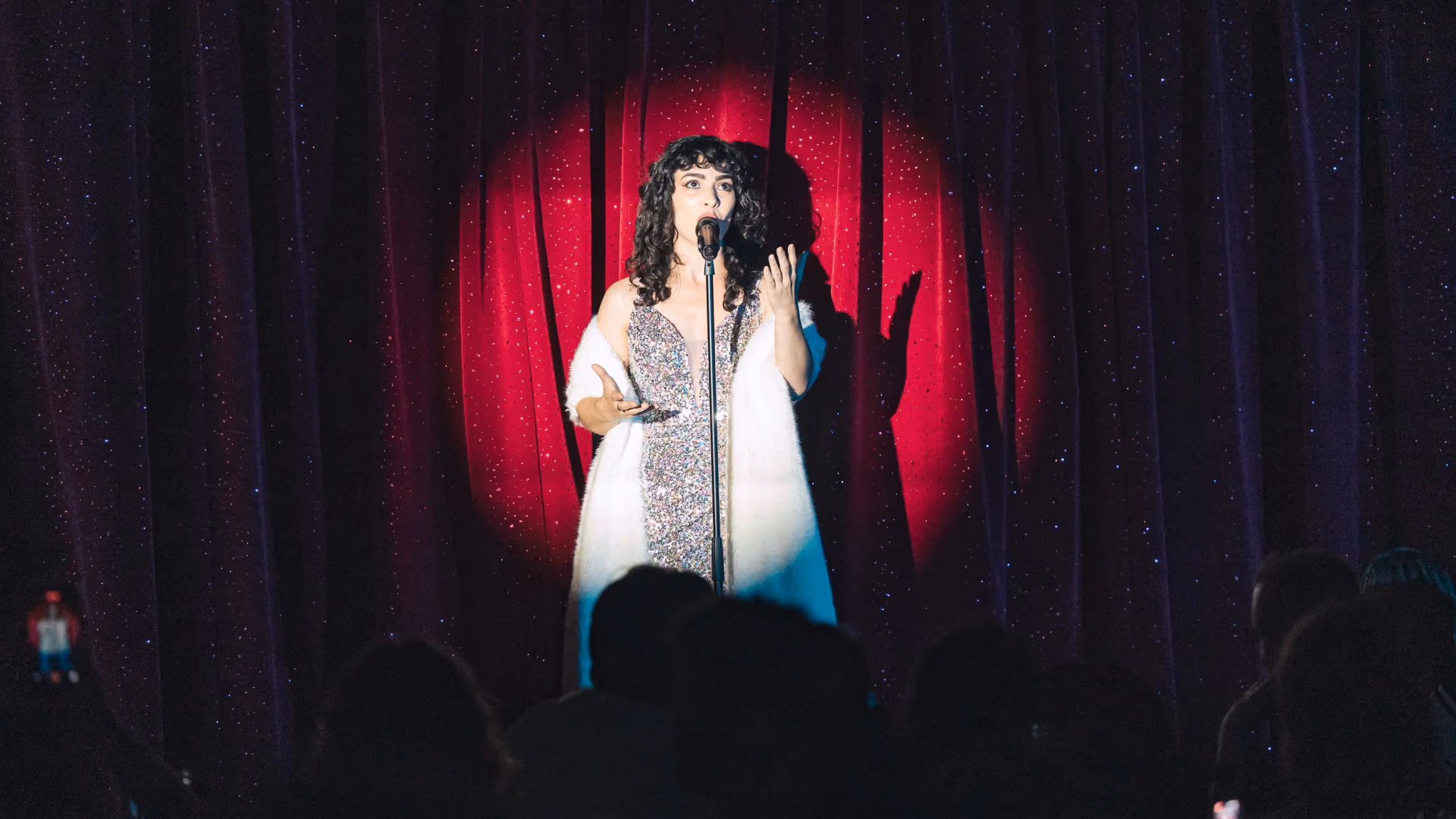 A person is singing into a microphone on stage in front of a red and black curtain, with spectators in silhouette in the foreground.