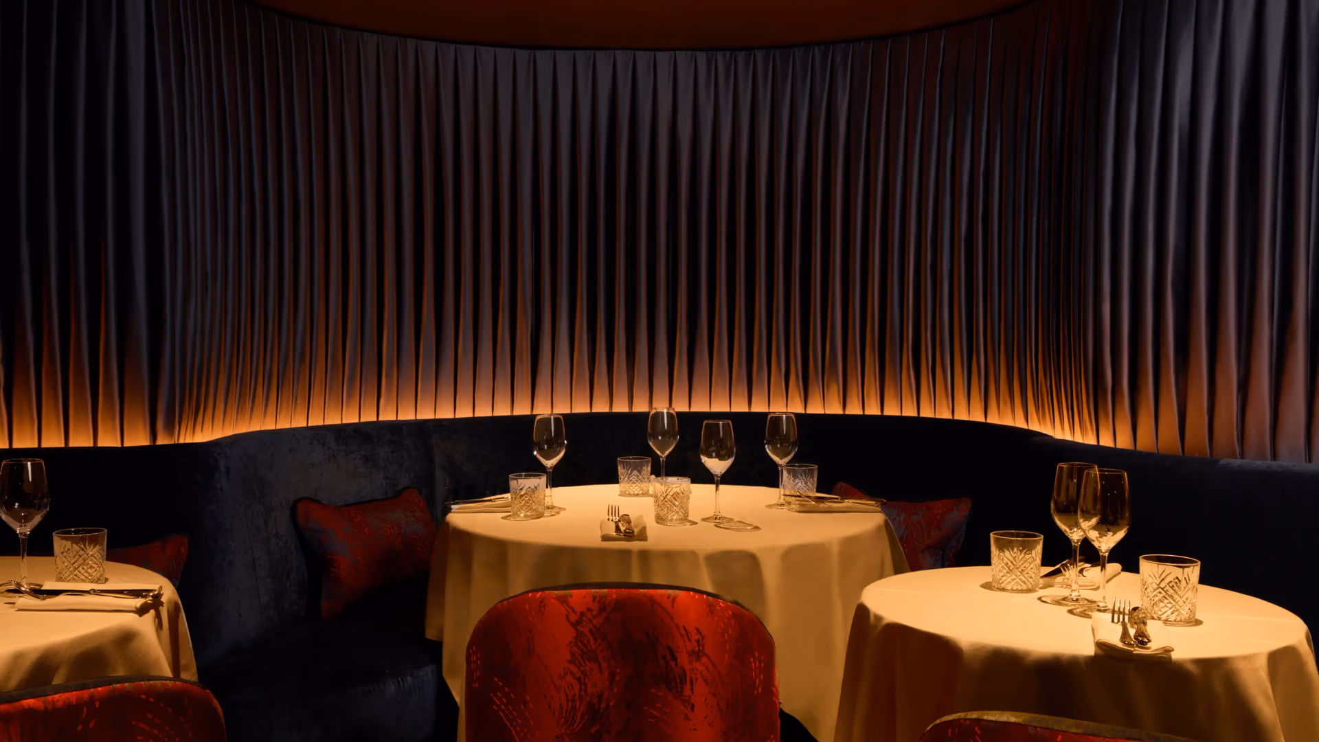 Une table ronde avec des verres et des couverts, entourée de chaises rouges, dans un restaurant à l'éclairage tamisé et aux rideaux sombres