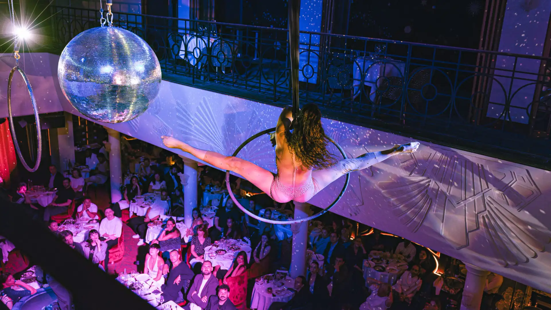 Une artiste en tenue scintillante fait un numéro de pole dance dans un club bondé, sous une boule à facettes et des lumières colorées