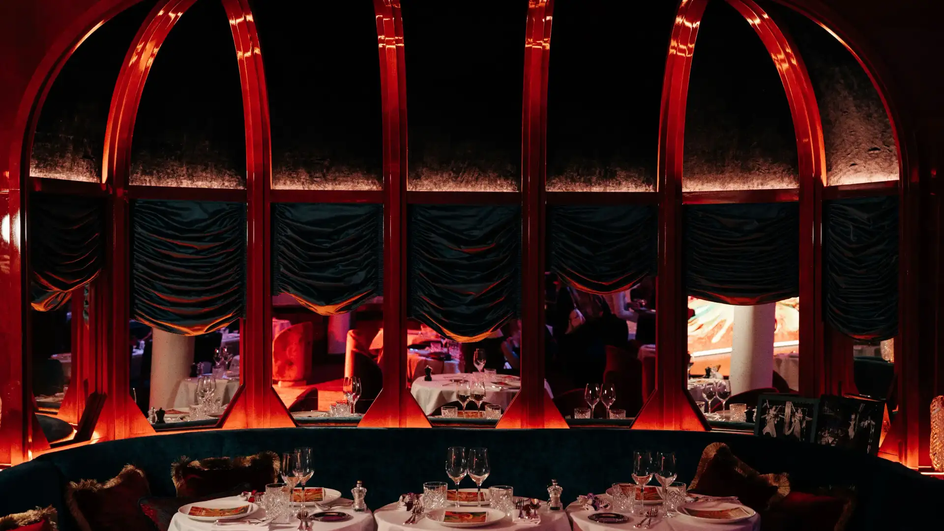 Three round tables with glasses and cutlery are arranged in front of a large red-arched window in a dark and elegant interior.